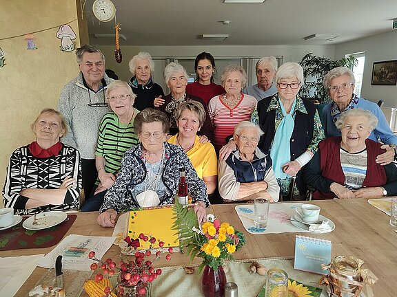 Eine Gruppe älterer Menschen sitzt im Tagesbetreuungszentrum mit den Betreuerinnen an einem Tisch auf dem sich selbst angefertigte Basteleien befinden.