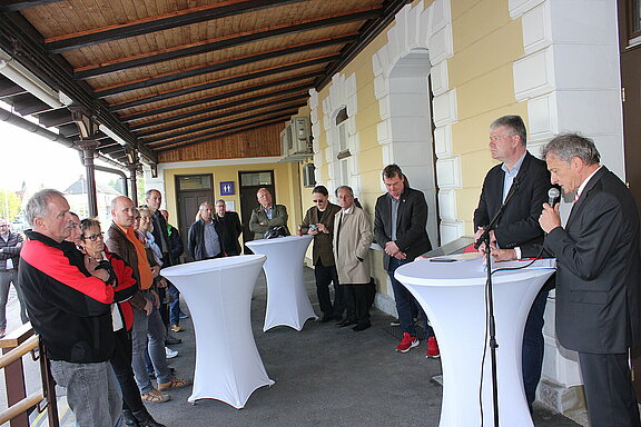 Eine Auswahl der Gäste, die der Enthüllungsfeier beiwohnten stehen unter dem Vordach des Bahnsteiges und lauschen den Worten von Dr. Johannes Kammerstätter.