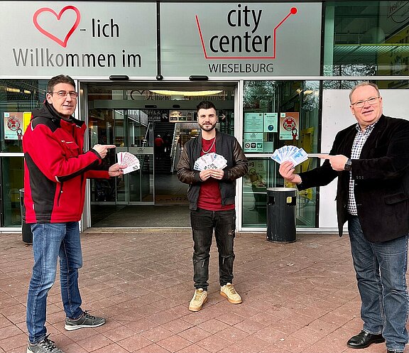 CityCenter Manager Dietmar Affengruber und der Vorsitzende des Stadtmarketings Wieselburg Josef Lechner übergeben dem Gewinner des 2. Preises Herrn Eduard Gashi seinen Gewinn. Sie stehen dabei im Freien vor dem Eingang zum CityCenter.