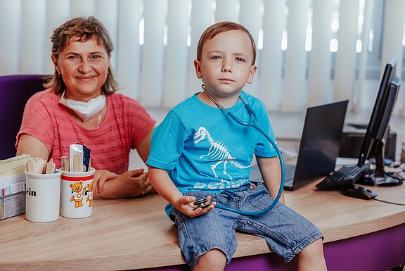 Porträt von Dr. Offenberger an ihrem Schreibtisch, auf dem ein Junge sitzt 