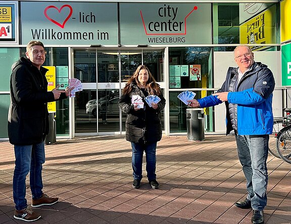 CityCenter Manager Dietmar Affengruber und der Vorsitzende des Stadtmarketings Wieselburg Josef Lechner übergeben der Gewinnerin des Hauptpreises Frau Regina Koppensteiner ihren Gewinn. Sie stehen dabei im Freien vor dem Eingang zum CityCenter.
